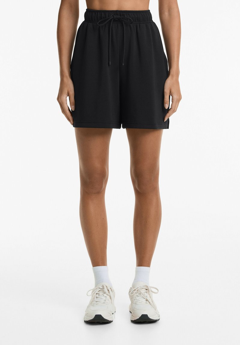 Shorts de sport noirs avec une taille élastique, un cordon de serrage et des poches latérales, portés avec des chaussettes blanches et des baskets claires.