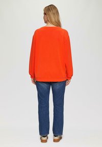 Helle orangefarbene übergroße Sweatshirt mit lässigem Schnitt, überschnittenen Schultern und gerippten Bündchen, kombiniert mit blauen geraden Jeans und beigen Sneakern.