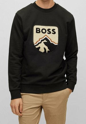 Sweat-shirt noir avec un patch texturé montrant un motif de montagne et le mot « BOSS », porté par une personne avec un pantalon beige et la main dans la poche.