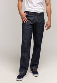 Mężczyzna w ciemnoniebieskich dżinsach, białej koszulce i granatowych trampkach z brązowymi sznurówkami, stojący na jednolitym beżowym tle.