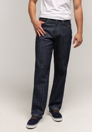 Mężczyzna w ciemnoniebieskich dżinsach, białej koszulce i granatowych trampkach z brązowymi sznurówkami, stojący na jednolitym beżowym tle.