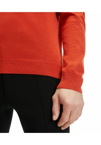 Roter Strickpullover mit langen Ärmeln, gerippten Bündchen und Saum, kombiniert mit schwarzen, enganliegenden Hosen. Weiche Textur und lässige Designdetails sichtbar.
