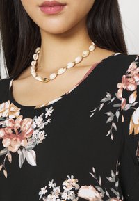 Collar de cuentas blancas con acentos dorados, llevado sobre una blusa negra con estampado floral que presenta flores rosas y blancas, con una textura suave y un escote en forma de barco.