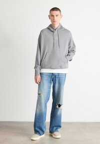 Siv hoodie z žepom spredaj, s šnurovanjem in kvadratnim logotipom. Nosi se čez belo plast. Počasen modri kavbojke, ohlapen kroj, veliki risi na kolenih. Bež čevlji.
