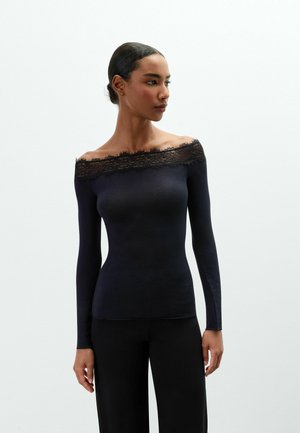 Femme aux cheveux foncés attachés en chignon, portant un haut noir à manches longues, épaules dénudées avec bordure en dentelle, et un pantalon noir, regardant vers sa gauche.