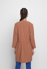 Brun långärmad kappa med elastisk midja, som har en avslappnad passform och mjukt tyg. Baksidan har en enkel rund halsringning.