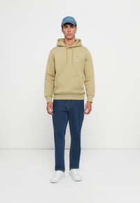 Ung mann iført beige hettegenser, blå jeans, hvite sneakers og lyseblå caps, står mot en enkel hvit bakgrunn.