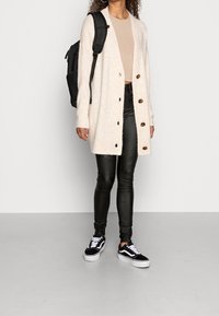 Kermanvärinen, pörröinen neuletakki, jossa ruskeat napit, beigellä topilla, yhdistetty kiiltäviin mustiin leggingsseihin ja mustiin lenkkareihin. Mukana musta reppu.