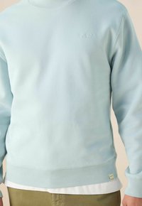 Sweatshirt bleu clair en tissu doux, avec un col rond, des poignets côtelés et un petit détail de texte en relief sur la poitrine.