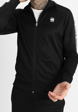Mann trägt eine schwarze Zip-Up-Jacke mit weißem G-Star RAW-Logo auf Brust und Ärmel, kombiniert mit schwarzen Hosen und weißem Unterhemd.