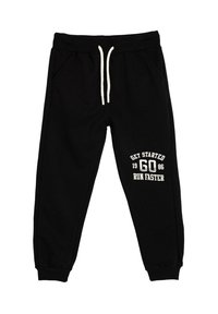 Zwarte sweatpants met een trekkoord in de taille, zijzakken en geribbelde manchetten, met witte tekst: "GA VAN START 1986 LOOP SNELLER."
