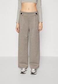 Breda byxor i houndstooth-mönster, tillverkade av ett texturerat tyg med framtill veck och svarta knapptyger. Bärs med silverfärgade sneakers.