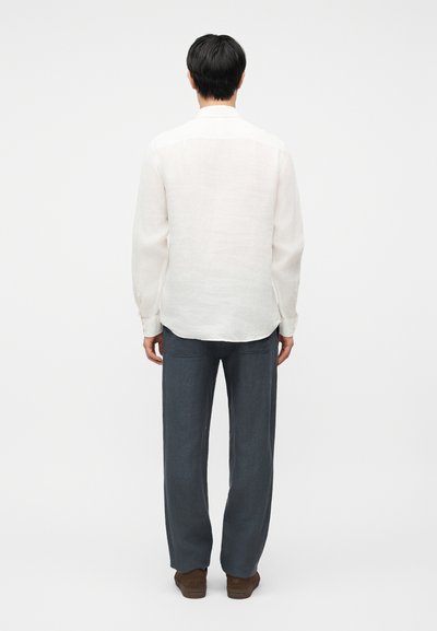 Άνδρας που στέκεται γυρισμένος από πίσω, φοράει λευκό πουκάμισο με μακριά μανίκια, σκούρο γκρι παντελόνι και καφέ παπούτσια σε απλό λευκό φόντο.