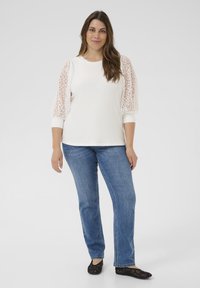 Top blanco con mangas de encaje, escote redondo y puños acanalados, combinado con vaqueros rectos azules y zapatos planos negros.