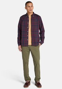 Camicia a quadri nei toni blu navy e borgogna con accentature gialle, indossata sopra una maglietta beige; abbinata a pantaloni verde oliva e scarpe marroni.