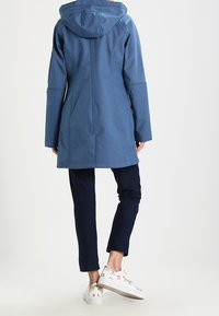 Blå vattentät parkas med huva, med en mjuk textur och långa ärmar. Styrelsen kombineras med marinblå byxor och vita sneakers med guldaccenter.