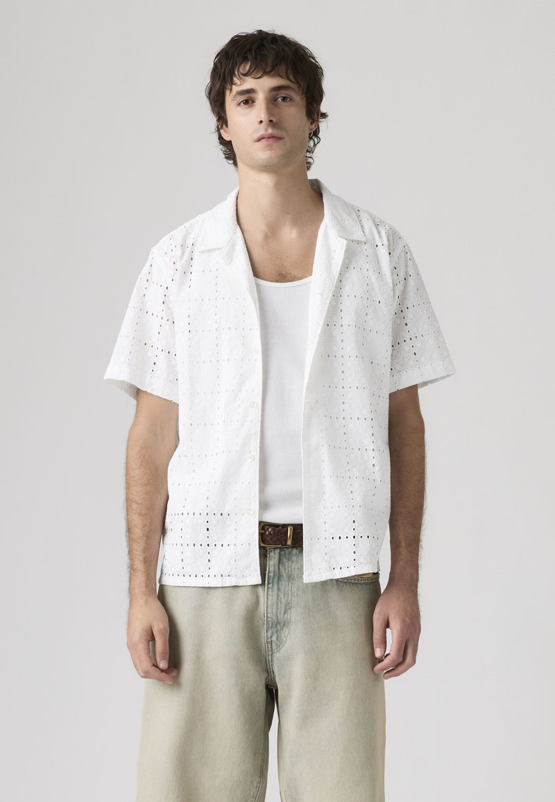 Junger Mann trägt ein weißes, gemustertes Kurzarmhemd über einem weißen Tanktop und hellbeigen Jeans vor einem schlichten grauen Hintergrund.