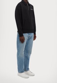 Svart zip-neck sweatshirt med känguruficka, ihopparad med ljusblå jeans och vita sneakers. Mjuk tyg, avslappnad passform, minimalistisk design.