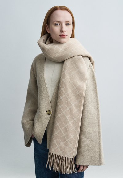 Μπεζ πλεκτό κασκόλ με γεωμετρικό μοτίβο, με φουντάκια στις άκρες, φορεμένο πάνω από ένα ανοιχτό πλεκτό τοπ και μια μπεζ ζακέτα. Λεία υφή και μαλακό υλικό.