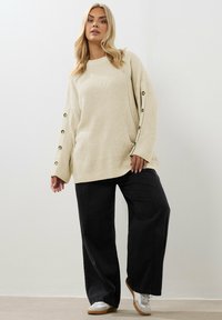 Sweter w kolorze kremowym, dziany, z szerokimi rękawami i złotymi guzikami, zestawiony z czarnymi spodniami o szerokich nogawkach i białymi trampkami.