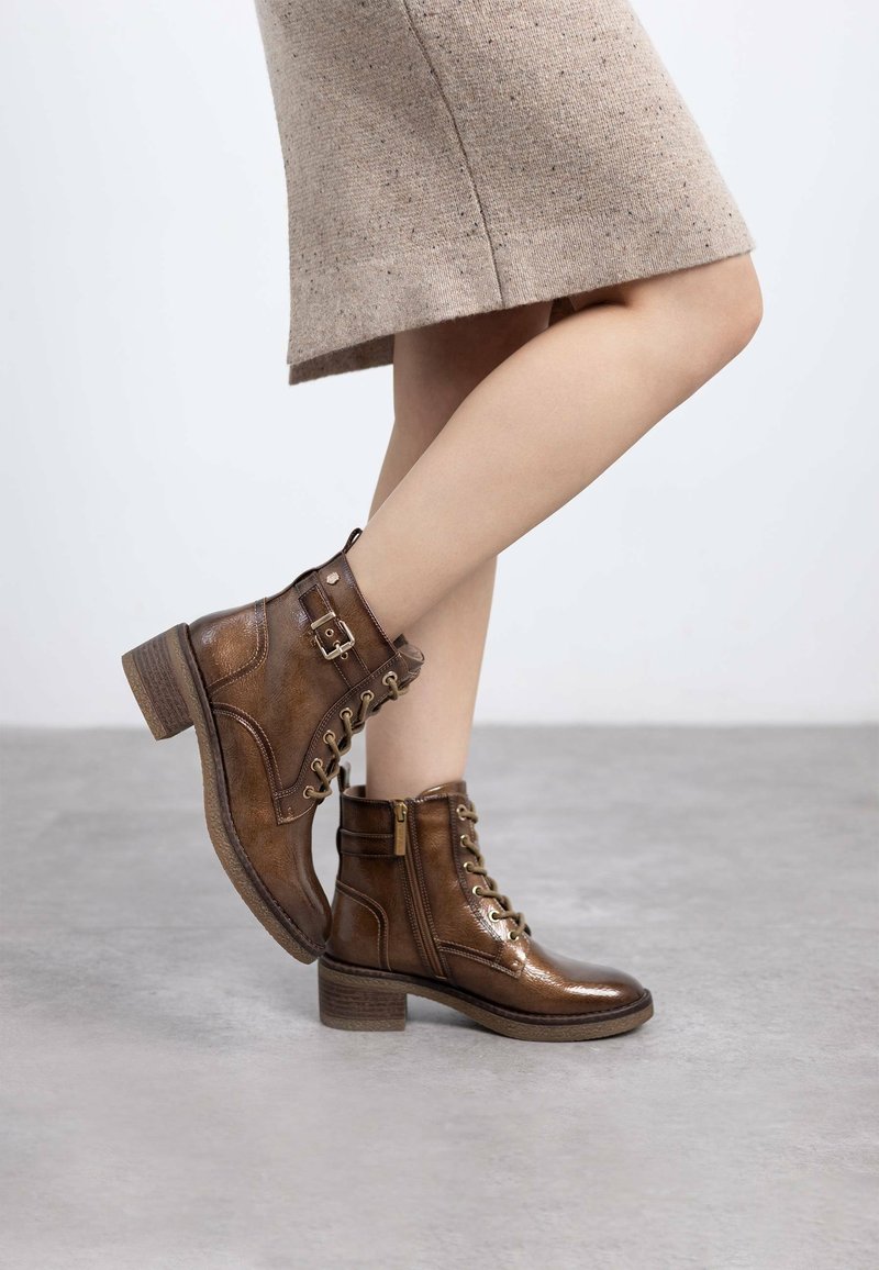 Botas de tobillo de cuero marrón con un acabado brillante, cordones en la parte delantera, cremallera lateral y un tacón apilado. El diseño presenta acentos metálicos y costuras.