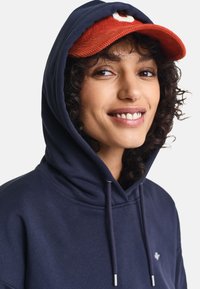 Navy blue hoodie with a drawstring, featuring a small logo on the chest. Paired with a red corduroy cap with a curved brim.