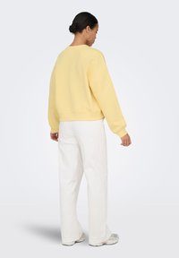 Vrouw met donker haar in een knot, draagt grote oorringen, een gele trui, witte broek met wijde pijpen en sneakers, kijkt van achteren tegen een effen achtergrond.