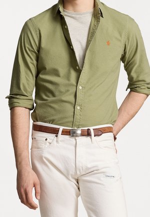 Homme portant une chemise boutonnée vert olive à manches retroussées par-dessus un t-shirt gris clair, un pantalon blanc et une ceinture en cuir marron.