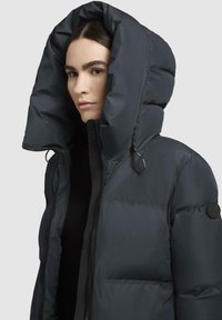 Giacca puffa blu navy con cappuccio oversize, dotata di chiusura con zip e logo applicato. Texture liscia e opaca con dettagli elasticizzati.