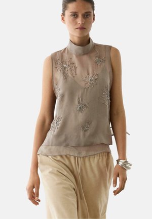 Kobieta nosząca bezrękawnik w kolorze taupe z przezroczystego materiału z koralikowymi wzorami wybuchających gwiazd, beżowe spodnie i srebrne bransoletki na prawym nadgarstku.