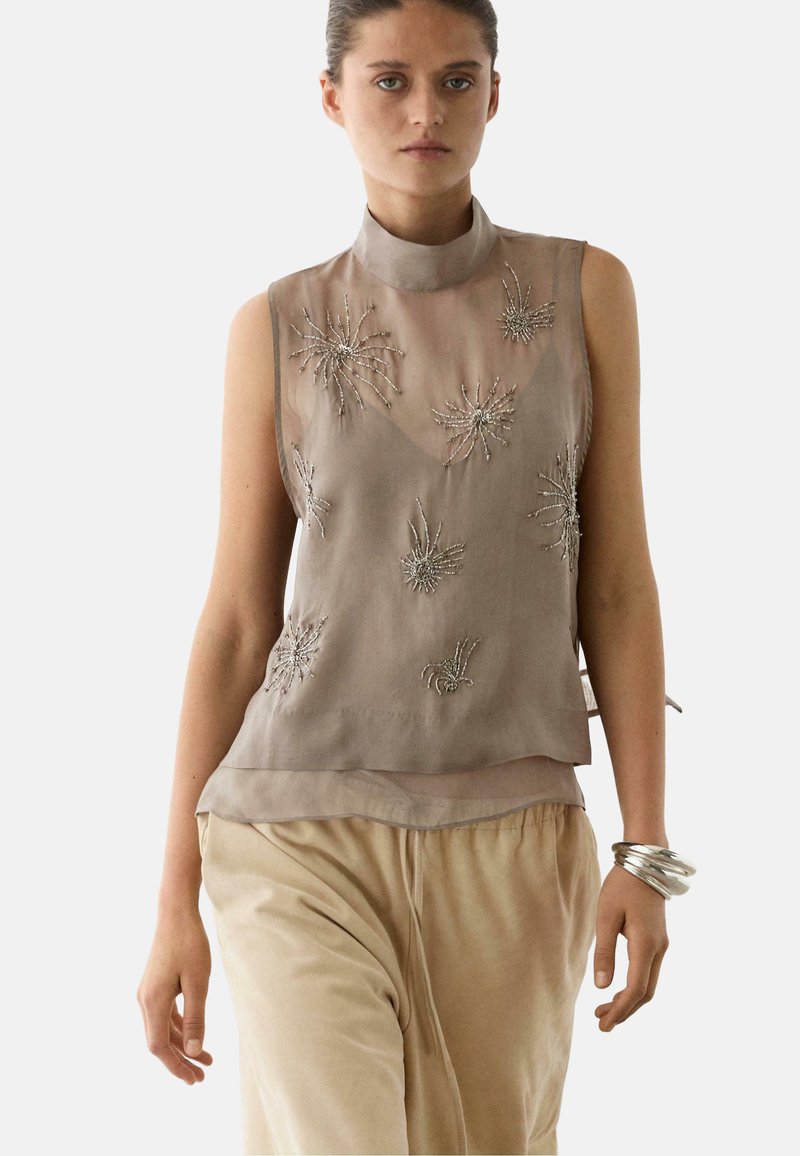 Vrouw draagt een mouwloos, taupekleurig, doorschijnend top met kralen in stervormige patronen, beige broek en zilveren armbanden om haar rechterpols.