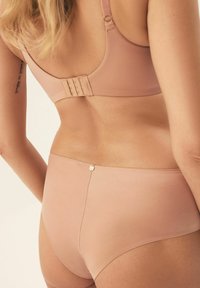 Beżowy biustonosz i majtki z wysokim stanem wykonane z gładkiego materiału. Biustonosz ma regulowane ramiączka i zapięcie na haftkę; majtki posiadają złoty guzik ozdobny.