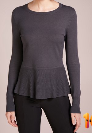 Vrouw die een donkergrijs gebreid shirt met lange mouwen draagt, met een geribbelde, uitlopende peplumzoom, en zwarte aansluitende broek, zichtbaar van nek tot heupen.