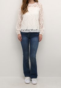 Top bianco in pizzo con maniche lunghe e orlo scallop, abbinato a jeans aderenti blu medio e sneakers bianche.
