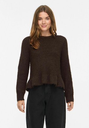 Vrouw met lichtbruin haar die een donkerbruine, pluizige trui en zwarte broek draagt, staand tegen een witte achtergrond.