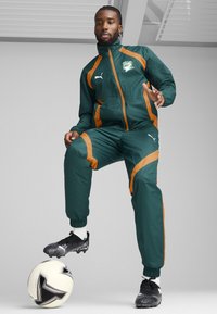 Green and orange track suit with zip jacket and elastic cuffs, paired with black soccer cleats, against a light gray background.