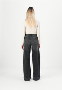 Kvinne med langt blondt hår iført en kremfarget langermet topp og høy i livet, vide mørkegrå jeans, står mot en hvit vegg.