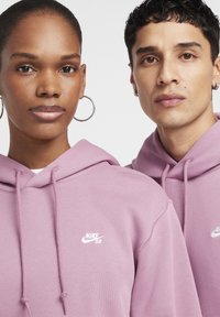 Dve osebi nosita ujemajoči se puloverji z kapuco v svetlo vijolični barvi. Puloverji imajo na prsih bel logotip Nike SB.