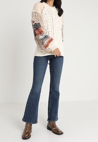 Pletený svetr v krémové barvě s vícebarevnými vzorovanými rukávy, kombinovaný s rozšířenými modrými džínami a kotníkovými botami s hadím potiskem.