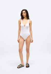 Γυναίκα με σγουρά μαλλιά, φορώντας λευκό κορμάκι χωρίς μανίκια και γκρι σανδάλια χωρίς λουριά, στέκεται μπροστά σε απλό λευκό φόντο.