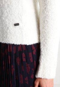 Pull blanc texturé à manches longues avec logo Napapijri sur la poitrine, associé à une jupe plissée noire ornée de motifs floraux rouge foncé.