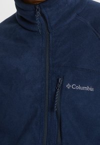 Giacca in pile blu navy con colletto alto, chiusura a zip completa e logo Columbia sul petto, realizzata in tessuto testurizzato per offrire calore.