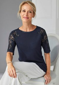 Marineblaue Bluse mit kurzen Ärmeln, die überdurchsichtige Spitzenakzente und perlengeschmückte Verzierungen am Ausschnitt verfügt. Weiches Material und lässige Passform.