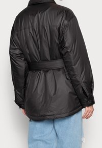 Chaqueta acolchada negra con cintura ceñida por un cinturón, que cuenta con cuello, mangas largas y puños abotonados, confeccionada en una tela lisa y brillante.