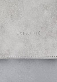 Sivá semišová peňaženka s vyrazeným logom "EXPATRIÉ", hladkým povrchom a viditeľným prešívaním na okraji.