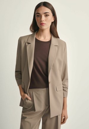 Kobieta w beżowej marynarce i spodniach, z ciemnobrązową bluzką, stojąca na jednolitym jasnym tle, z ręką w kieszeni.