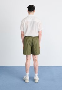 Camisa de manga curta, com riscas claras e colarinho aberto, combinada com calções elásticos verde-oliva. Ténis brancos e meias completam o visual.