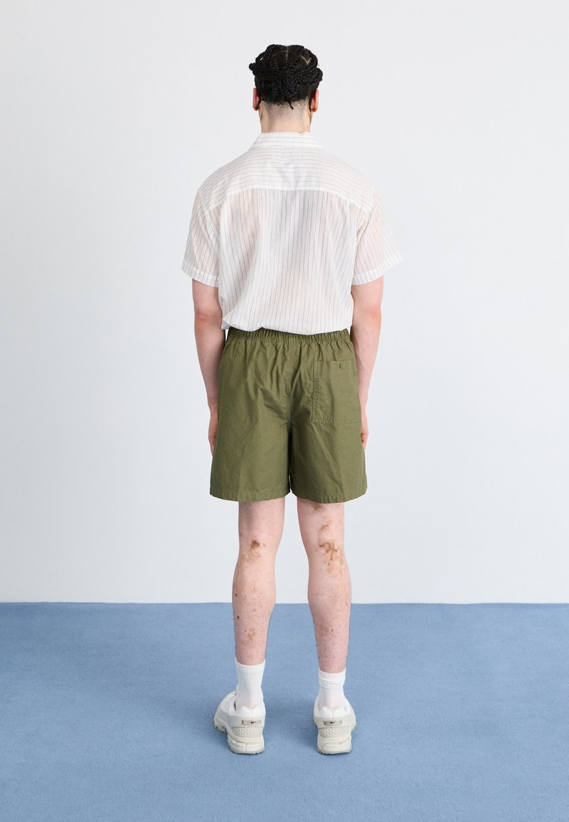 Kurzarmhemd mit feinen Streifen und offenem Kragen, kombiniert mit olivgrünen Shorts mit elastischem Bund. Weiße Sneakers und Socken runden den Look ab.