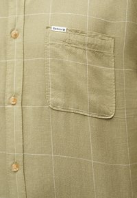 Ανοιχτό πράσινο πουκάμισο ελιάς με λευκό καρό μοτίβο, ξύλινα κουμπιά και τσέπη στο στήθος με την επιγραφή "Barbour" στην αριστερή πλευρά.