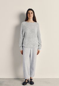 Woman standing against beige wall wearing a light gray fuzzy sweater, matching joggers, and black woven sandals.